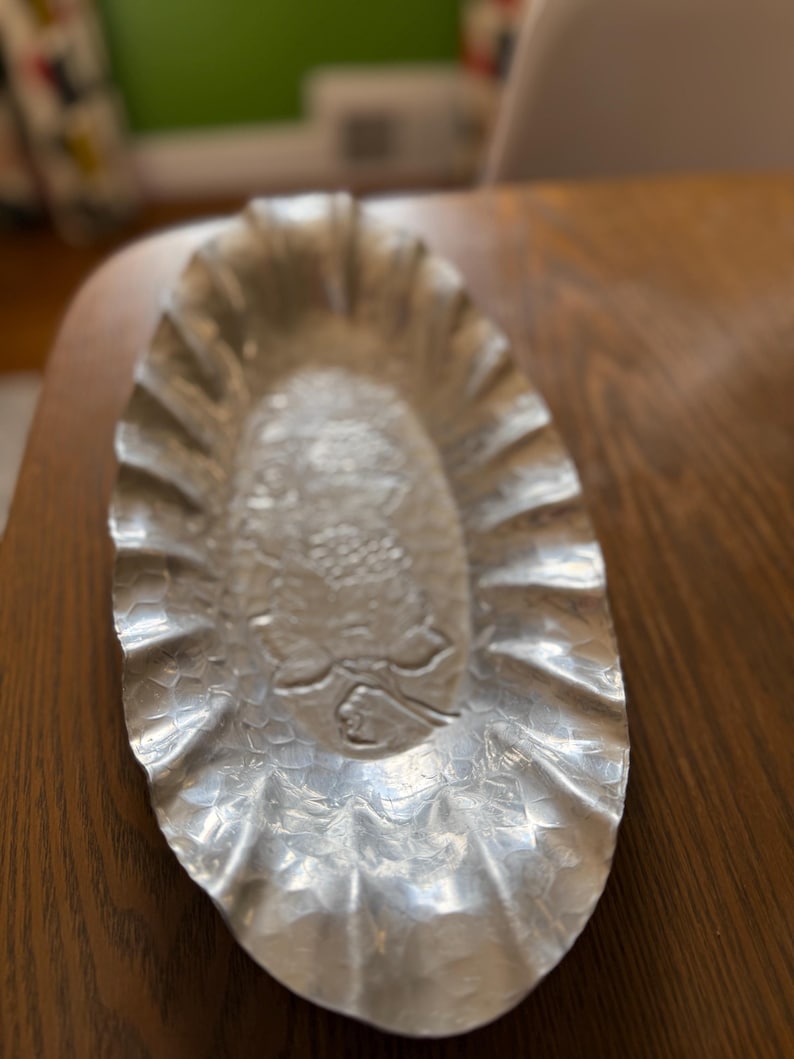 K&ouml;nnte beinhalten: Silberfarbene, ovale Servierschale mit strukturierter Oberfl&auml;che und ger&uuml;schtem Rand. Die Mitte der Schale weist ein detailliertes gepr&auml;gtes Design auf. Die Schale steht auf einem Holztisch.