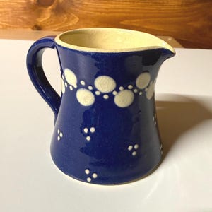 May include: A blue ceramic pitcher with a handle and spout. The pitcher is decorated with white polka dots and a cream-colored interior. The pitcher is likely used for serving beverages.