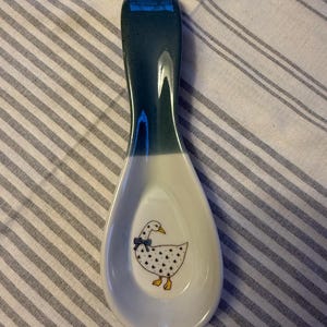 May include: White ceramic spoon rest with a teal handle and a goose illustration. The goose has black spots and a blue bow. The spoon rest is designed to hold cooking utensils and protect surfaces from spills.