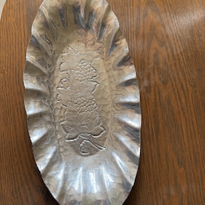 K&ouml;nnte beinhalten: Ovale, geh&auml;mmertes Metallserviergeschirr mit ger&uuml;schtem Rand. Die Mitte zeigt ein Weinreben-Design. Das Geschirr ist silberfarben und steht auf einer Holzoberfl&auml;che. Ein dekoratives St&uuml;ck zum Servieren von Snacks oder zum Pr&auml;sentieren von Gegenst&auml;nden.