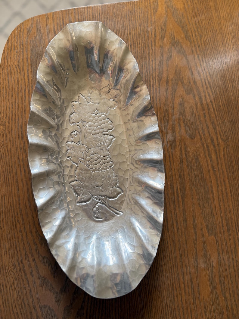 K&ouml;nnte beinhalten: Ovale, geh&auml;mmertes Metallserviergeschirr mit ger&uuml;schtem Rand. Die Mitte zeigt ein Weinreben-Design. Das Geschirr ist silberfarben und steht auf einer Holzoberfl&auml;che. Ein dekoratives St&uuml;ck zum Servieren von Snacks oder zum Pr&auml;sentieren von Gegenst&auml;nden.