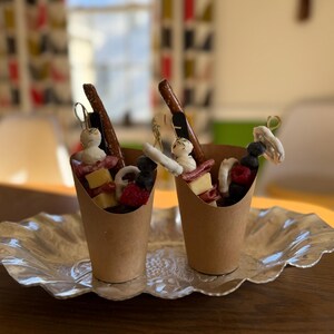 K&ouml;nnte beinhalten: Zwei Papiert&uuml;ten mit Charcuterie-Zutaten, darunter K&auml;se, Beeren und Brezeln, stehen auf einer dekorativen Silberplatte. Die Becher sind auf einem Holztisch angeordnet, was auf einen Snack oder eine Vorspeise hindeutet.