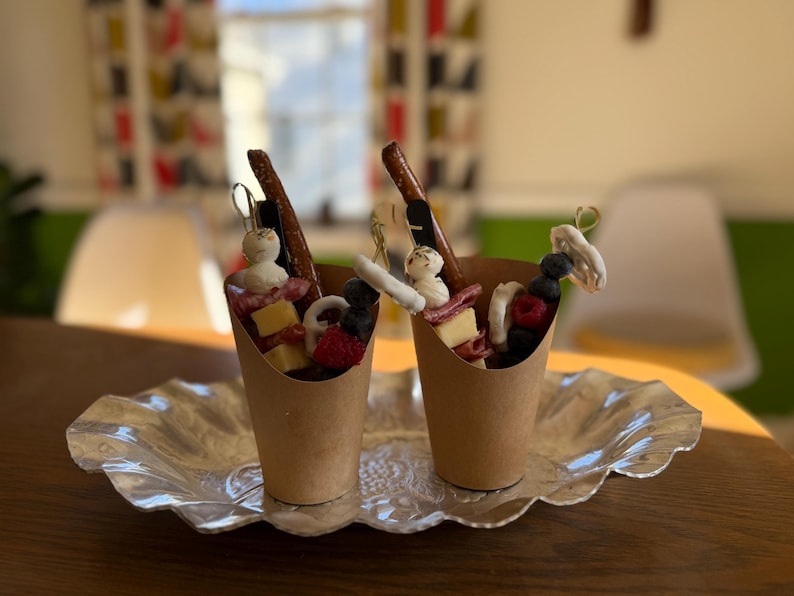 K&ouml;nnte beinhalten: Zwei Papiert&uuml;ten mit Charcuterie-Zutaten, darunter K&auml;se, Beeren und Brezeln, stehen auf einer dekorativen Silberplatte. Die Becher sind auf einem Holztisch angeordnet, was auf einen Snack oder eine Vorspeise hindeutet.