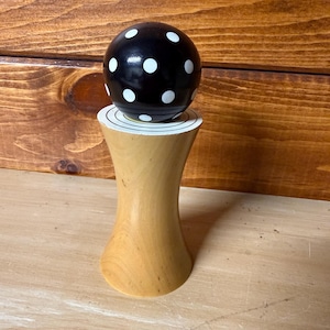 May include: A wooden bottle stopper with a black, polka-dotted sphere top. The stopper has a light wood base with a flared design. The base is approximately 4 inches tall.