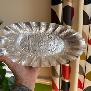 K&ouml;nnte beinhalten: Ovale, silberfarbene Servierplatte mit geh&auml;mmertem Muster und ger&uuml;schtem Rand. Die Mitte der Platte zeigt ein erhabenes Design aus Trauben und Bl&auml;ttern. Die Platte wird von einer Person gehalten.