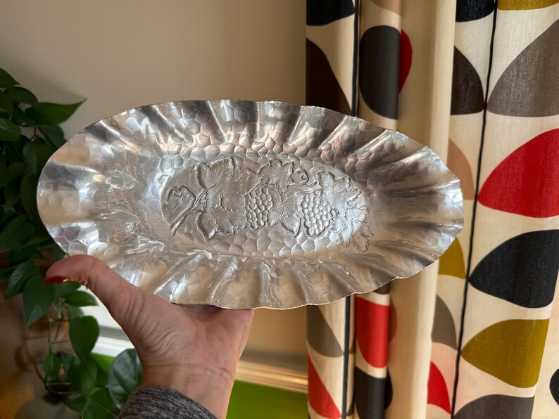 K&ouml;nnte beinhalten: Ovale, silberfarbene Servierplatte mit geh&auml;mmertem Muster und ger&uuml;schtem Rand. Die Mitte der Platte zeigt ein erhabenes Design aus Trauben und Bl&auml;ttern. Die Platte wird von einer Person gehalten.