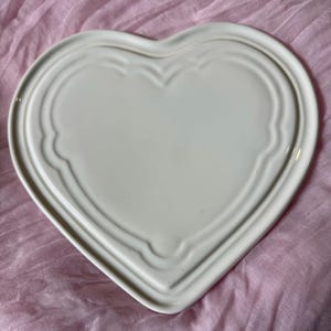 May include: A heart-shaped, off-white ceramic plate. The plate features a raised, scalloped border that follows the heart's outline. The plate is smooth and has a glossy finish. The plate is designed for serving food.