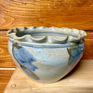 May include: A handmade ceramic bowl with a ruffled rim. The bowl features a light blue and beige color scheme with blue abstract floral designs. The bowl is sitting on a wooden surface.