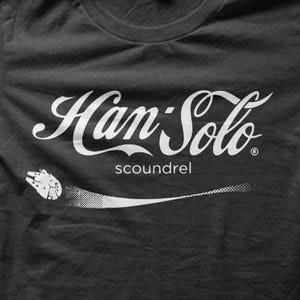 May include: Black t-shirt with a white graphic that reads "Han Solo" in a stylized font, with the word "scoundrel" below. A white image of the Millennium Falcon is depicted in flight, with a dotted trail.