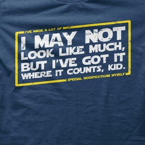 Może przedstawiać: Granatowa koszulka z białym i żółtym prostokątnym nadrukiem. Na nadruku widnieje napis: "I MAY NOT LOOK LIKE MUCH, BUT I'VE GOT IT WHERE IT COUNTS, KID." Obecny jest również tekst "I'VE MADE A LOT OF" i "SPECIAL MODIFICATIONS MYSELF".