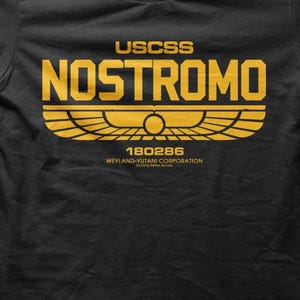 May include: Black t-shirt featuring the yellow text "USCSS NOSTROMO". Below the text is a yellow graphic of a stylized bird with outstretched wings. The shirt also features the text "180286" and "WEYLAND-YUTANI CORPORATION".