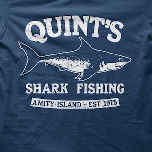 May include: Navy blue t-shirt with a white graphic. The graphic reads "QUINT'S SHARK FISHING" with a shark illustration and "AMITY ISLAND EST. 1975". The shirt is a crew neck style.