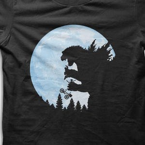 Könnte beinhalten: Schwarzes T-Shirt mit einer Silhouette eines Monsters, das ein kleines Fahrzeug vor einem blauen Mond hält. Das Design beinhaltet einen Wald am unteren Rand. Die Grafik ist eine Parodie einer berühmten Filmszene.