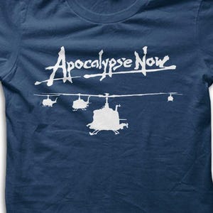 May include: Navy blue t-shirt featuring the text "Apocalypse Now" in white script above a line of white helicopter silhouettes. The design is centered on the front of the shirt.