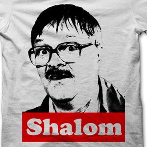 May include: White t-shirt featuring a black and white graphic of a man with glasses and a mustache. The word "Shalom" is printed in white on a red rectangle at the bottom of the shirt. The shirt is made of a soft material.