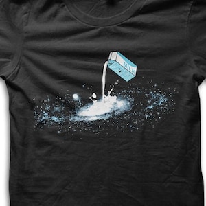 Könnte beinhalten: Schwarzes T-Shirt mit einem verspielten Design eines Milchkartons, der Milch in einen galaxieartigen Spritzer aus Weiß und Blau gießt. Die Grafik ist mittig platziert und bietet einen auffälligen und fantasievollen Look.