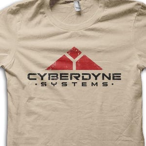 Könnte beinhalten: Beigefarbenes T-Shirt mit dem Text "CYBERDYNE SYSTEMS" in Schwarz und einem roten, verblassten Dreieckslogo. Das T-Shirt hat einen Rundhalsausschnitt und kurze Ärmel. Das Design ist auf der Vorderseite aufgedruckt.