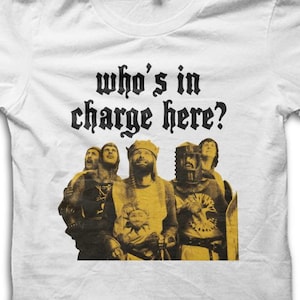 Pode incluir: Camiseta branca com um gráfico de um grupo de pessoas em trajes medievais. O texto "who's in charge here?" é impresso acima do gráfico em uma fonte gótica. O gráfico é em tons de amarelo e preto.