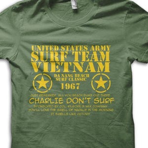 Könnte beinhalten: Olivgrünes T-Shirt mit gelbem Text. Der Text lautet "UNITED STATES ARMY SURF TEAM VIETNAM 1967" mit Sterngrafiken. Darunter steht "CHARLIE DON'T SURF" und weitere Sätze.