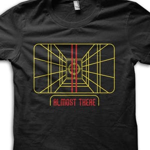 Puede incluir: Camiseta negra con un diseño gráfico retro. El diseño incluye una cuadrícula amarilla, dos líneas verticales rojas y el texto "ALMOST THERE" en rojo. El gráfico tiene una estética de ciencia ficción o videojuego.