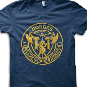 May include: Navy blue t-shirt featuring a distressed yellow circular graphic with the text "NORGES" and "TROLLSIKKERHETSTJENESTE". The graphic includes axes and the letters "TSI".