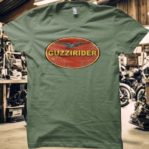 May include: Olive green t-shirt with a distressed oval design. The design features the text "GUZZIRIDER" in yellow, a red background, and a black bird graphic. The shirt has a crew neck and short sleeves.