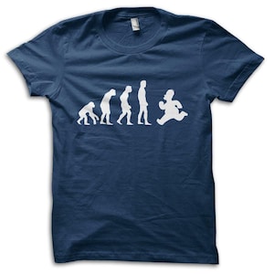 Può includere: T-shirt blu navy con grafica bianca raffigurante l'evoluzione dell'uomo, che culmina con un personaggio dei cartoni animati in corsa. Il design mostra la progressione dalla scimmia all'uomo, con la figura finale in posizione di corsa. Classica maglietta girocollo.