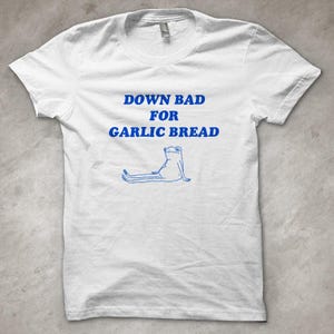 Puede incluir: Camiseta blanca con el texto azul "DOWN BAD FOR GARLIC BREAD" y una ilustración azul de una rana. La rana está sentada con las piernas estiradas. La camiseta tiene cuello redondo y mangas cortas.