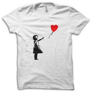 May include: White t-shirt featuring a black silhouette of a young person reaching for a red heart-shaped balloon. The design is a graphic illustration, with the balloon floating away. The shirt is a classic crew neck style.