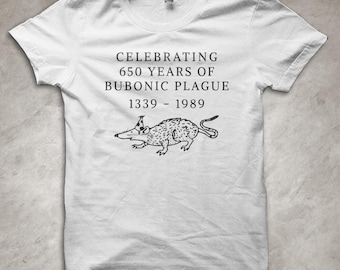 Peut inclure: T-shirt blanc avec l'inscription "CELEBRATING 650 YEARS OF BUBONIC PLAGUE 1339 - 1989" et une illustration en noir et blanc d'un rat. Le t-shirt a des manches courtes et un col rond. Design humoristique.