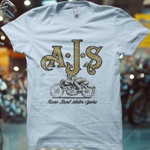 Pode incluir: Camiseta azul clara com um gráfico dourado desgastado. O gráfico inclui as letras "AJS" acima de uma ilustração de motocicleta, com o texto "Race-Bred Motor Cycles" abaixo. A camiseta tem gola redonda e mangas curtas.