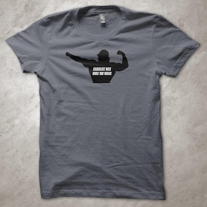 Peut inclure: T-shirt gris avec une silhouette noire d'un bodybuilder en train de se muscler. Le texte "CHOCOLATE MILK GIVES YOU WINGS" est imprimé sur le torse. Le t-shirt a un col rond et des manches courtes.