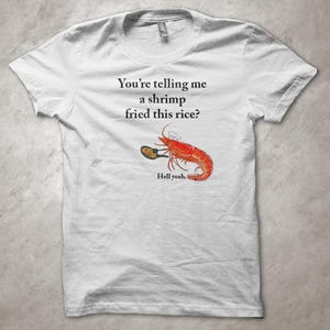 Può includere: T-shirt bianco con grafica umoristica. Il design presenta un gamberetto dei cartoni animati che tiene in mano una padella di riso fritto, con il testo "You're telling me a shrimp fried this rice?" e "Hell yeah."
