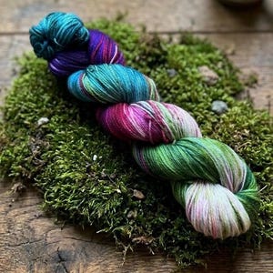 May include: A skein of colorful yarn in shades of teal, purple, pink, and green, resting on a bed of green moss. The yarn is twisted and shows a gradient of colors. The background is a rustic wooden surface.