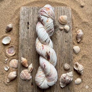 May include: A skein of white yarn with red and brown speckles, arranged on a weathered wooden plank. The yarn is surrounded by various seashells on a sandy surface, creating a coastal theme. The yarn is a twisted, rope-like structure.