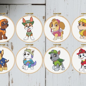 May include: Eight cross-stitch embroidery hoops featuring cartoon dog characters from the Paw Patrol series. Each hoop has a different dog character, with varying colors and outfits, set against a white background.