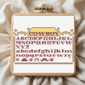 May include: Cross-stitch sampler with the word "COWBOY" in a yellow and brown font. The sampler includes the alphabet in uppercase and lowercase, plus cowboy-themed icons. The sampler is in a wooden hoop.