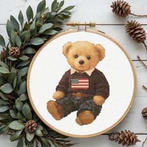 May include: Cross-stitch embroidery of a teddy bear wearing a brown sweater with an American flag patch and plaid pants. The bear is in a wooden embroidery hoop. Decorative greenery and pinecones surround the hoop.