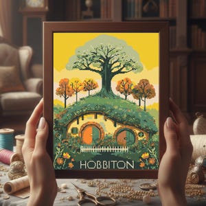 May include: A framed cross-stitch depicting a Hobbiton scene with a large tree, rolling hills, and two round hobbit doors. The artwork features a yellow sky and the word "HOBBITON" at the bottom. The frame is brown.