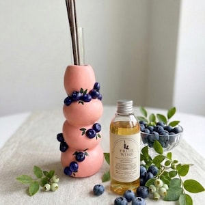 Puede incluir: Un difusor de cerámica rosa apilado con detalles de arándanos, un vial de vidrio con varillas y una botella de perfume "Fruit Wine". Arándanos frescos y vegetación se disponen alrededor de los objetos.