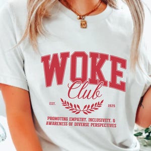 May include: White t-shirt with red text that reads "WOKE Club Est. 1925 Promoting Empathy. Inclusivity. & Awareness of Diverse Perspectives".