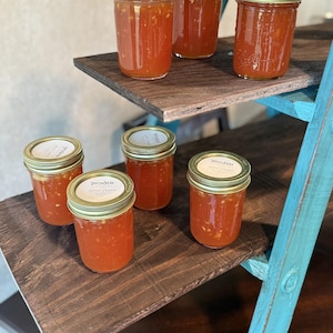 May include: Six glass jars filled with a reddish-orange preserve, each sealed with a gold lid. The jars are arranged on a dark wooden shelf supported by a turquoise ladder-like structure. The labels read "Tropical".