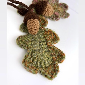 May include: A handmade crochet necklace featuring a green, brown, and orange oak leaf pendant with two brown acorns. The leaf pendant is detailed with textured stitching, and the acorns have a textured cap. The necklace is on a brown cord.