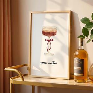 Può includere: Una stampa incorniciata con un'illustrazione di espresso martini. L'opera raffigura un bicchiere da cocktail con un fiocco rosso, riempito con un liquido marrone a strati. Le parole "espresso martini" sono scritte sotto il bicchiere in corsivo. Sono visibili anche una bottiglia di liquore e un vaso con foglie verdi.