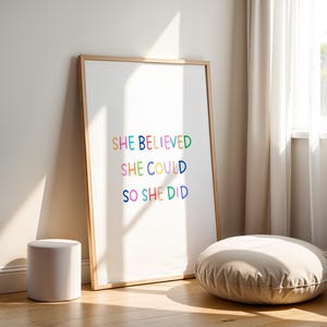 May include: A framed print with the text "SHE BELIEVED SHE COULD SO SHE DID" in colorful letters. The print has a light wood frame and is leaning against a wall. A small white cylindrical stool and a round floor cushion are in the foreground.