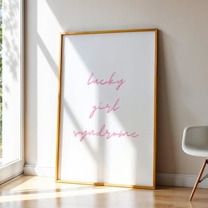 Pode incluir: Uma impressão de arte emoldurada com as palavras "lucky girl syndrome" em script rosa. A obra de arte com moldura dourada está encostada a uma parede branca, ao lado de uma cadeira branca com pernas de madeira. A sala tem um chão de madeira e luz natural.