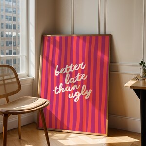 Può includere: Una stampa artistica incorniciata con uno sfondo a strisce rosa e rosse vibranti. Il testo bianco recita "better late than ugly". L'opera d'arte è appoggiata a un muro vicino a una finestra e a una sedia di legno.