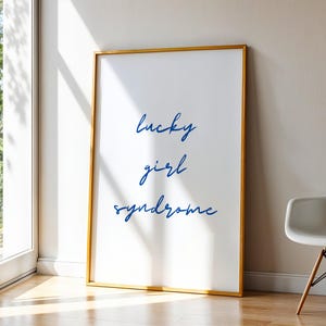 Può includere: Una stampa artistica incorniciata con le parole "lucky girl syndrome" in blu su sfondo bianco. La cornice è dorata e la stampa è appoggiata a un muro vicino a una finestra e a una sedia bianca.