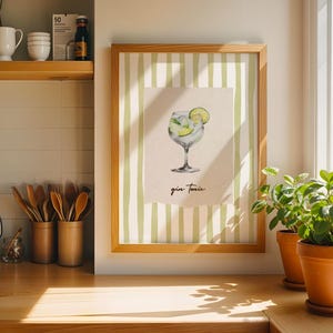 Può includere: Una stampa incorniciata con un'illustrazione di un cocktail gin tonic. L'opera presenta un gin tonic con lime, su uno sfondo a strisce verdi e bianche. La cornice è in legno chiaro.