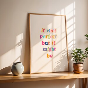 Può includere: Una stampa incorniciata con le parole "it isn't perfect but it might be" in testo colorato. La stampa è esposta su una mensola di legno con un vaso grigio e una pianta in vaso. La cornice è di colore marrone chiaro.